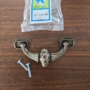 Puede incluir: Tirador de armario de latón envejecido con un diseño central acanalado y dos tornillos de montaje. El tirador va acompañado de un paquete de plástico con el texto "Amerock Limited, Meaford, Ontario".