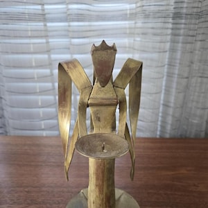 Op de afbeelding: Een vintage messing engelenkaarsenhouder. De engelenfiguur heeft uitgespreide vleugels en een kroon. De kaarsenhouder is rond met een centrale punt om een kaars in te plaatsen. Het messing heeft een warme, verouderde patina.