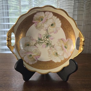May include: An antique serving dish with a scalloped edge and gold trim. The dish features a floral design with white and pink flowers. The dish is displayed on a black stand.