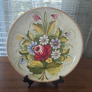 Op de afbeelding: Een decoratieve keramische plaat met een handgeschilderd bloemmotief. De plaat toont een centraal boeket van rode, witte, blauwe en paarse bloemen met groene bladeren. Een gouden rand omlijnt de rand van de plaat, die op een zwarte standaard wordt getoond.
