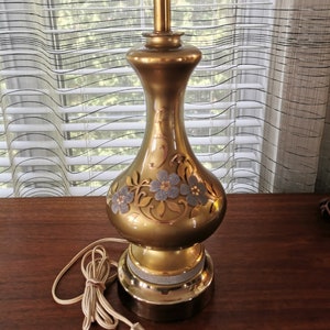 Peut inclure: Une lampe de table dorée avec un motif floral. La lampe a un cordon blanc et une prise blanche.