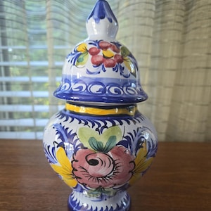 Puede incluir: Un jarrón de cerámica pintado a mano con tapa. El jarrón es blanco con diseños florales azules, amarillos y rosas. La tapa está rematada con un pináculo azul. El jarrón mide aproximadamente 20 cm de altura.