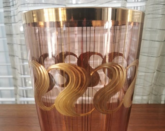 May include: A tall, clear glass vase with a pink hue and gold accents. The vase has a gold rim and a pattern of gold swirls around the middle.
