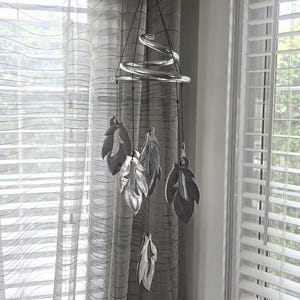 May include: A silver-toned wind chime with a spiral design and leaf-shaped pendants. The chime hangs from a black string and is suspended in front of a window with sheer curtains and blinds.