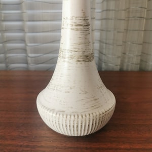 Puede incluir: Un jarrón de cerámica blanco con un acabado texturizado y desgastado y una base acanalada. El jarrón tiene un cuello estrecho y un cuerpo ancho y redondeado.