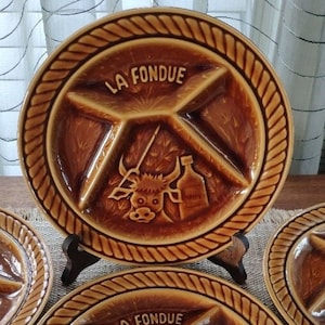 May include: Two brown ceramic fondue plates with a rope design border. Each plate has four sections and features a bull and a bottle with the text "LA FONDUE" in the center.