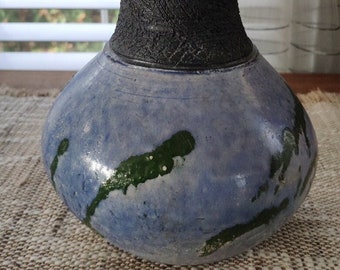May include: A handmade ceramic vase with a textured black neck and a rounded, blue body. The blue surface has green accents. The vase is a decorative item, suitable for displaying flowers or as a standalone art piece.
