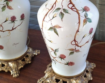 Może przedstawiać: Para białych ceramicznych lamp stołowych z złotymi akcentami. Lampy mają kwiatowy wzór z brązowymi gałęziami, zielonymi liśćmi i czerwonymi jagodami. Podstawy są ozdobne i mają złote wykończenie.