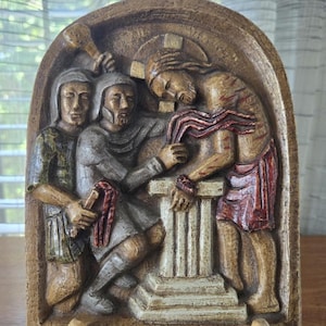 May include: A carved wooden relief depicting a religious scene with figures in robes. The relief is in shades of brown, beige, and red, mounted on a wooden easel. The figures are positioned around a central pillar.