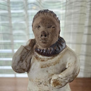 May include: A ceramic figurine of a person wearing a white top with a brown collar and brown boots. The figure has a neutral expression and is standing. The sculpture is on a wooden surface.
