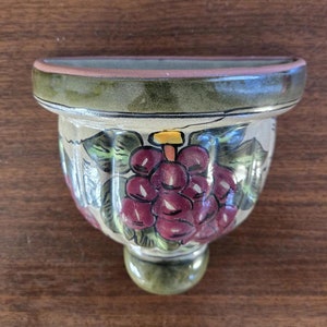 Puede incluir: Un macetero de pared de cerámica con un esmalte verde, blanco y marrón. El macetero está decorado con un diseño pintado de uvas y hojas.