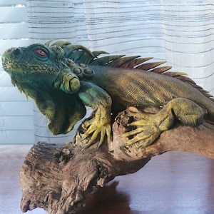 Puede incluir: Una figurita de iguana verde y amarilla posada sobre una rama de madera flotante desgastada. La iguana tiene un cuerpo texturizado, escamoso y una cola larga.