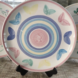 May include: A white ceramic plate with a pink rim and a colourful design. The plate features concentric circles in blue, yellow, and pink, with butterfly-shaped accents in various pastel colours. The plate is displayed on a black stand.