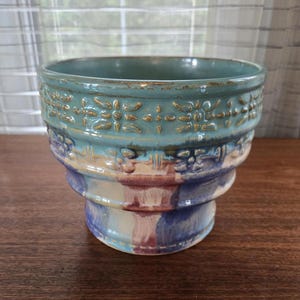 May include: A handcrafted ceramic planter with a teal exterior and a decorative floral pattern. The planter has a wide opening and a pedestal base, with bands of colour in blue, purple, and cream. Suitable for indoor or outdoor use.
