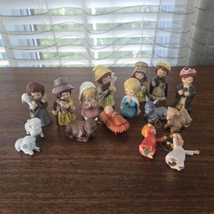 Może przedstawiać: Kolekcja małych ceramicznych figurek szopki bożonarodzeniowej, w tym postacie ludzi, niemowlę w żłobie, osioł, wielbłąd i owce. Figurki są pomalowane na różne kolory, a niektóre mają złote sznurki umożliwiające ich zawieszenie.