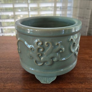 Puede incluir: Un pequeño macetero redondo de cerámica verde con un diseño floral decorativo y tres patas.
