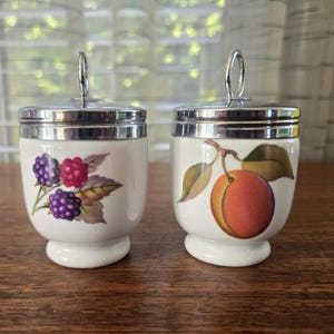 May include: Two white ceramic egg cups with silver lids. One cup features a design of blackberries, the other a peach. The egg cups are on a wooden surface.
