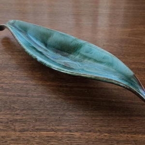 May include: A handcrafted, leaf-shaped ceramic dish with a glossy, teal glaze. The elongated, curved design features a raised edge and a subtle, swirling pattern within the teal colour. The dish is displayed on a dark wooden surface.