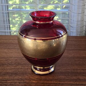 Peut inclure: Un vase en verre rouge rubis avec une bande dorée et un motif doré complexe autour du milieu. Le vase a un corps rond et une lèvre évasée.