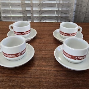 May include: Four white espresso cups and saucers. Each cup features a red band with the text "Cimbalino". The cups and saucers are arranged on a dark wooden surface.