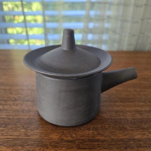 Puede incluir: Una pequeña tetera de cerámica gris oscuro con tapa y boquilla. La tetera está sobre una superficie de madera. La tapa tiene un pequeño asa en la parte superior. La boquilla se extiende desde el lateral de la olla.