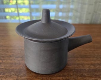 Op de afbeelding: Een kleine, donkergrijze keramische theepot met een deksel en een tuit. De theepot staat op een houten oppervlak. Het deksel heeft een klein handvat aan de bovenkant. De tuit steekt uit de zijkant van de pot.