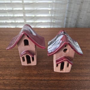 Puede incluir: Dos casas de cerámica en miniatura con tejados rojos y paredes de color beige. Los tejados tienen un acabado brillante, uno de ellos con un patrón veteado blanco y negro. Cada casa tiene una pequeña puerta y una ventana.