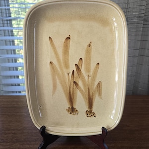 May include: Rectangular ceramic serving platter with rounded corners. The beige platter features a brown floral design. The platter is displayed on a dark brown wooden stand. The background includes a window and sheer curtains.