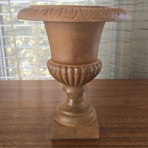 Puede incluir: Una urna decorativa marrón con una parte superior ancha y plana y una base detallada y acanalada. La urna tiene una base cuadrada y una superficie texturizada. La urna está hecha de un material duradero y es adecuada para uso en interiores o exteriores.