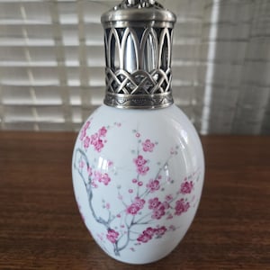 Op de afbeelding: Een witte keramische oliediffuser met een zilveren metalen bovenkant. De diffuser is versierd met een bloemenontwerp van roze bloesems en grijze takken. De metalen bovenkant heeft een ingewikkeld, decoratief patroon.