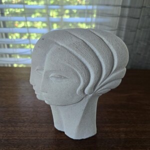 May include: A light gray, sculpted bust of a woman's head. The sculpture features detailed facial features and stylized hair. The bust is made of a textured material and sits on a wooden surface.