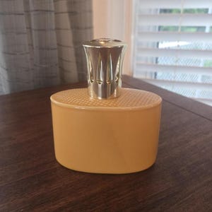 Könnte beinhalten: Eine ovale Duftlampe aus hellorangefarbener Keramik mit einem glatten, einfarbigen Körper und einer strukturierten Oberseite. Die Lampe verfügt über einen metallischen, silberfarbenen Aufsatz mit vertikalen Schlitzen, der oben aufsitzt.