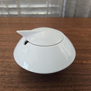 Könnte beinhalten: Eine weiße Zuckerdose aus Keramik mit Deckel. Der Deckel hat einen kleinen, spitzen Griff. Die Schüssel ist glatt und glänzend, mit einer abgerundeten Form. Die Zuckerdose steht auf einer Holzoberfläche.