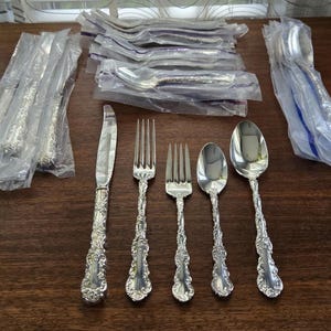 May include: A set of silver cutlery with ornate handles, including a knife, fork, and spoon. The cutlery is packaged in plastic bags.