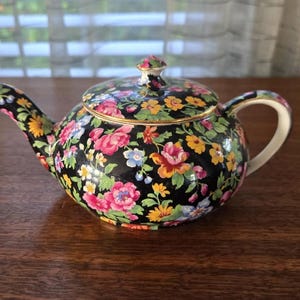 Puede incluir: Tetera negra adornada con un vibrante estampado floral en rosa, amarillo y azul. La tetera tiene un borde dorado y una tapa a juego. El asa y el pico son blancos con detalles florales. Un artículo decorativo para los amantes del té.
