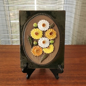 Puede incluir: Un azulejo de cerámica marrón con un diseño ovalado en relieve que presenta un grupo de flores amarillas, blancas y naranjas. El azulejo está enmarcado con un borde verde oscuro y tiene un borde con cuentas.