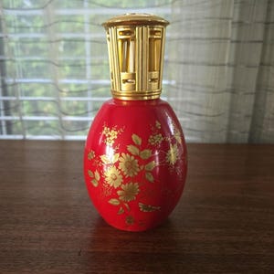 May include: A red glass fragrance lamp with a gold floral pattern featuring various blossoms and leaves. The lamp has a metallic gold-coloured vented cap at the top, which is cylindrical with rectangular cutouts around its perimeter.