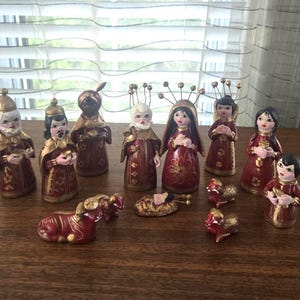 May include: A nativity set featuring eleven hand-painted figurines in deep red and gold tones. The collection includes standing figures with wire halos, a central infant in a manger, and several small animal figures, all arranged on a flat surface.