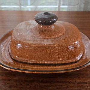 以下が含まれることがあります： 蓋付きの茶色の陶器製のバター入れ。皿は濃い茶色の縁取りと斑点のある釉薬が施されています。