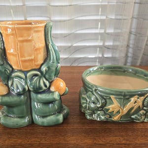 Puede incluir: Dos macetas de cerámica verde con figuras de elefantes. Una maceta es un jarrón alto con un diseño de bambú amarillo. La otra maceta es un cuenco redondo con un diseño de bambú y figuras de elefantes.