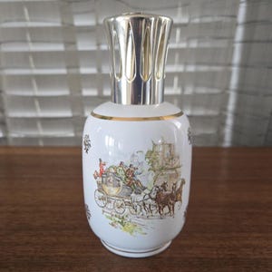 Op de afbeelding: Witte keramische geurlamp met een zilverkleurige bovenkant en een gouden rand. De lamp heeft een gedetailleerde illustratie van een paardenkoets met mensen. Decoratieve zwarte bloemaccenten zijn ook aanwezig.