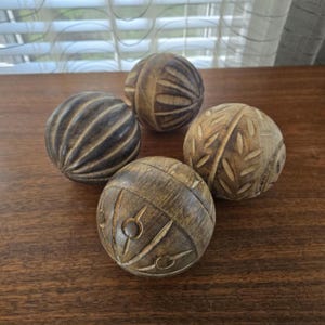 Puede incluir: Cuatro objetos esféricos de madera con texturas talladas. Cada esfera presenta un patrón distinto, incluyendo ranuras verticales, formas de hojas y motivos circulares. La madera muestra tonos marrones naturales con vetas variadas.