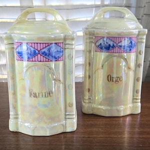 May include: Two iridescent ceramic canisters with handles. Each has a decorative band with blue and pink designs. One reads "Farine" and the other "Orge" in gold lettering. The canisters are on a wooden surface.