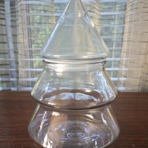 May include: A clear glass jar shaped like a Christmas tree. The jar has a conical lid and a two-tiered base. The jar is sitting on a wooden surface, with a window and sheer curtains in the background.