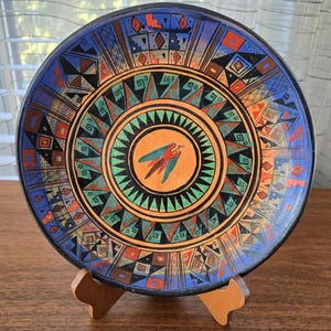 Puede incluir: Un plato decorativo de cerámica con un diseño geométrico vibrante. El plato presenta un círculo central naranja con una ilustración de un pájaro, rodeado de anillos concéntricos de patrones coloridos en azul, rojo, verde y negro. El plato se exhibe sobre un soporte de madera.