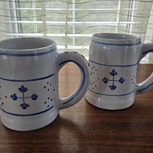 May include: Two white ceramic mugs with blue and white designs. The mugs have a cross-shaped design with blue accents.