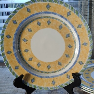 May include: A set of four yellow, blue, and green ceramic dinner plates with a diamond pattern. The plates are stacked on a black stand.