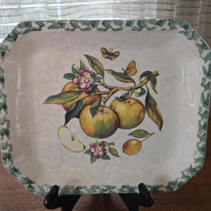 Puede incluir: Bandeja rectangular con fondo crema y borde verde. La bandeja presenta una ilustración detallada de manzanas, flores y mariposas. Las manzanas son amarillas y verdes, y las flores son rosas.