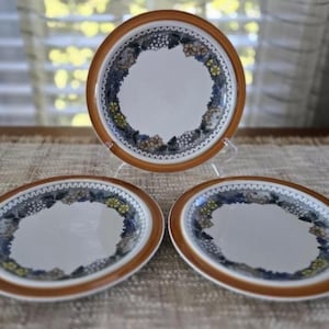 Puede incluir: Tres platos de cerámica con centro blanco y borde marrón. Los platos presentan un borde decorativo con diseños florales y de uvas azules, amarillos y blancos. Los platos están dispuestos sobre una superficie neutra.