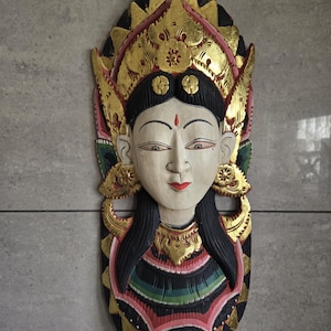 May include: A hand-carved wooden wall mask featuring a stylized face with a white complexion, black hair, and red lips. The mask is adorned with intricate gold-painted headwear and decorative elements, accented with green, pink, and black details.